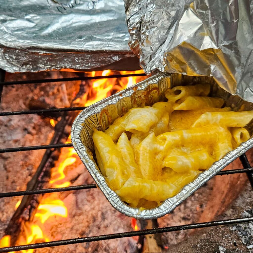 camping mac and cheese cooked over the campfire in aluminum pans in this easy make ahead camping dinner